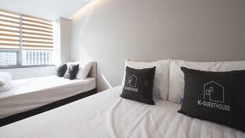 A modern hotel room with a bed, two pillows with 'K-GUESTHOUSE' branding, a window with blinds, and a city view.