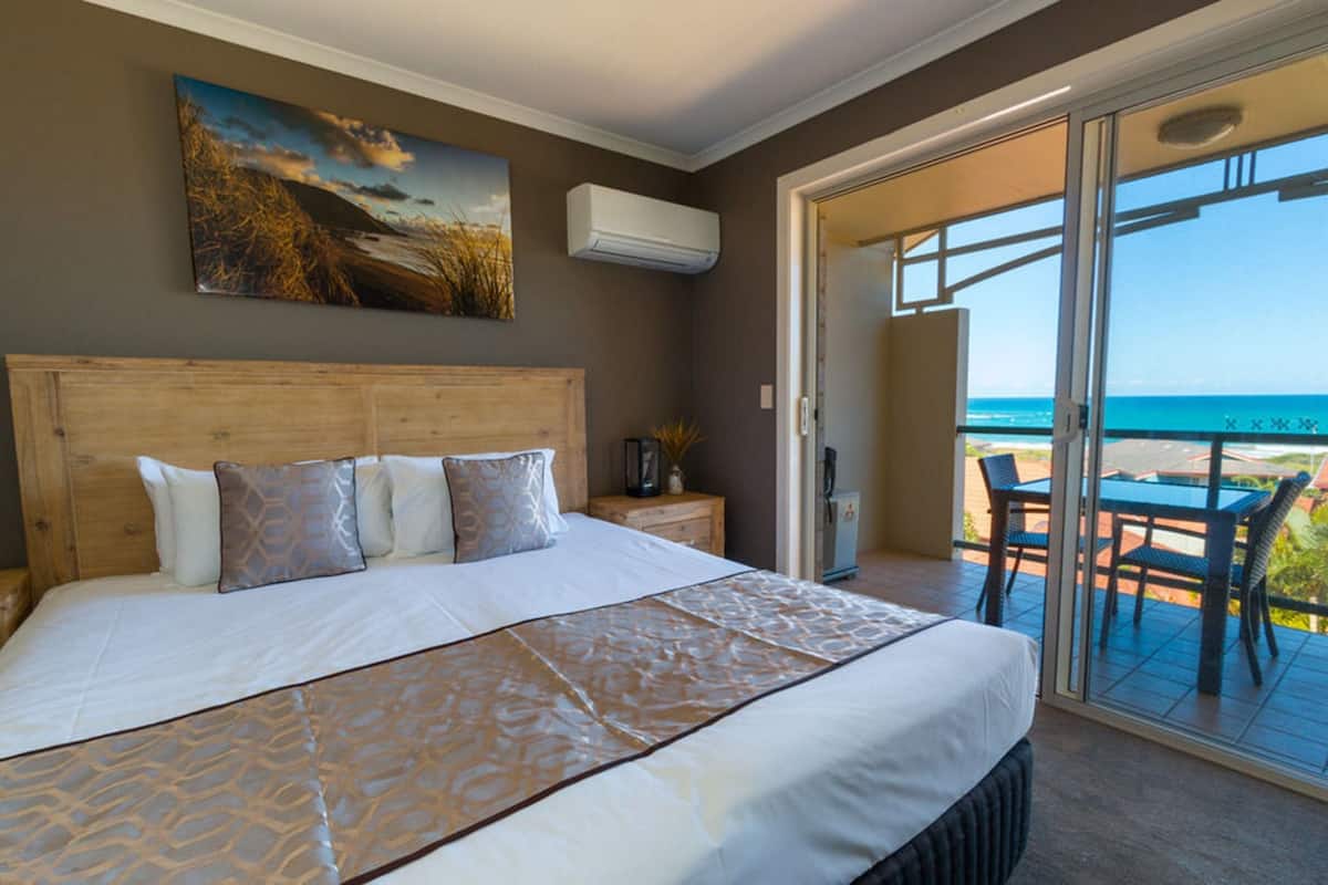 A bedroom with a large bed, a painting above the bed, an air conditioner on the wall, and a balcony with a table and chairs.