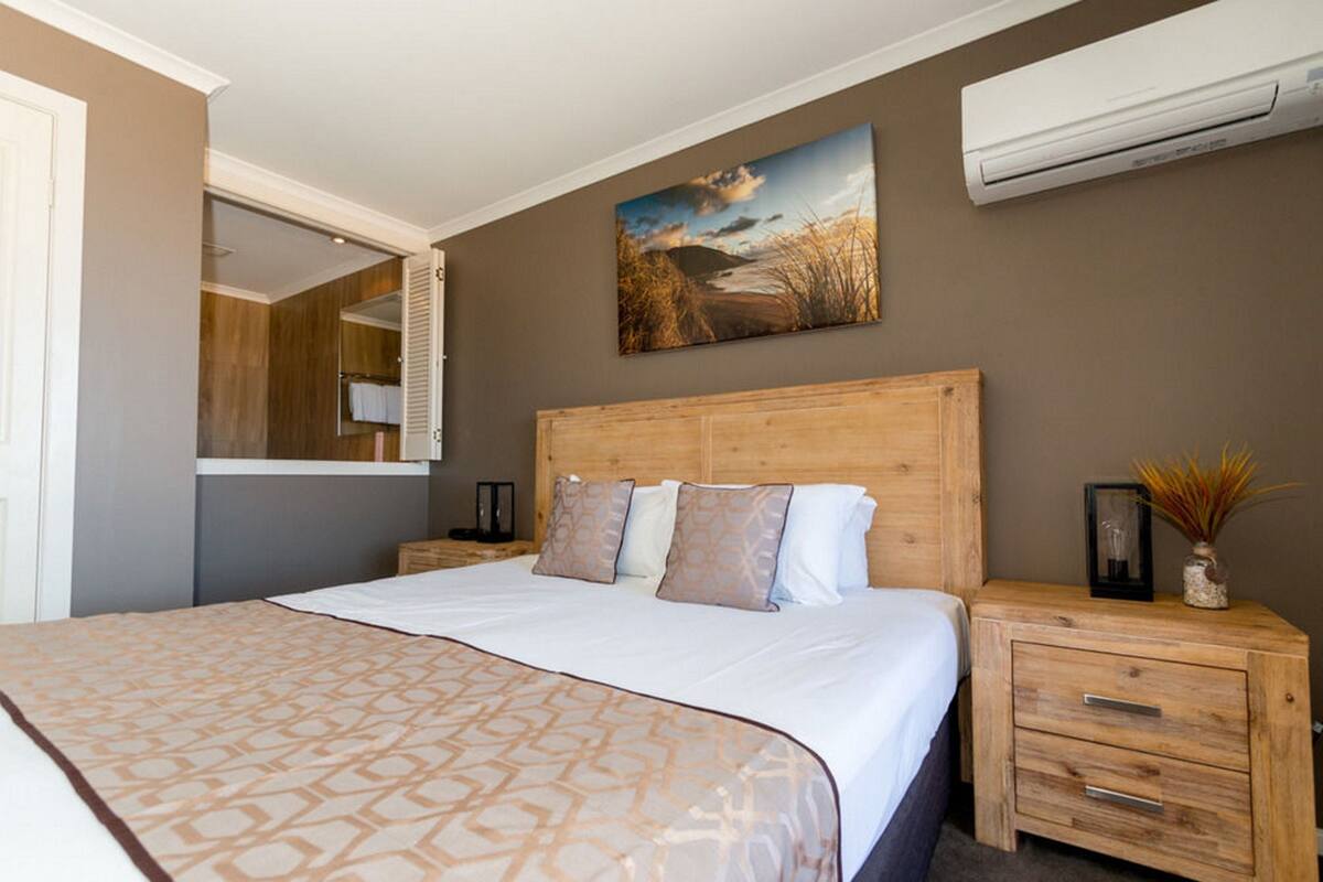 A bedroom with a large bed, wooden headboard, a nightstand with drawers, a mirror, and an air conditioning unit on the wall.