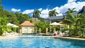Une piscine entourée de chaises longues et de parasols, bordée de palmiers et d’un bâtiment au toit rouge.