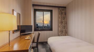 A hotel room with a bed, a desk with a lamp, a television, and a window with a city view.