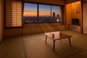 Een kamer met tatami vloerbedekking, een verschuifbaar shoji scherm, een televisie en een uitzicht op een stadsbeeld in de schemering.