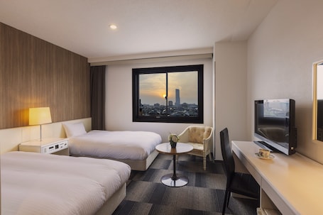 A hotel room with two beds, a desk with a TV, a chair, a small table with a flower vase, and a window with a cityscape view.. City Hotel Airport in Prince