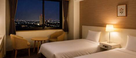 A hotel room with two beds, a small table, a chair, a nightstand with a lamp, and a window with a city view.