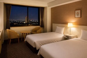 Een hotelkamer met twee bedden, een kleine tafel, een stoel, een nachtkastje met een lamp en een raam met uitzicht op de stad.