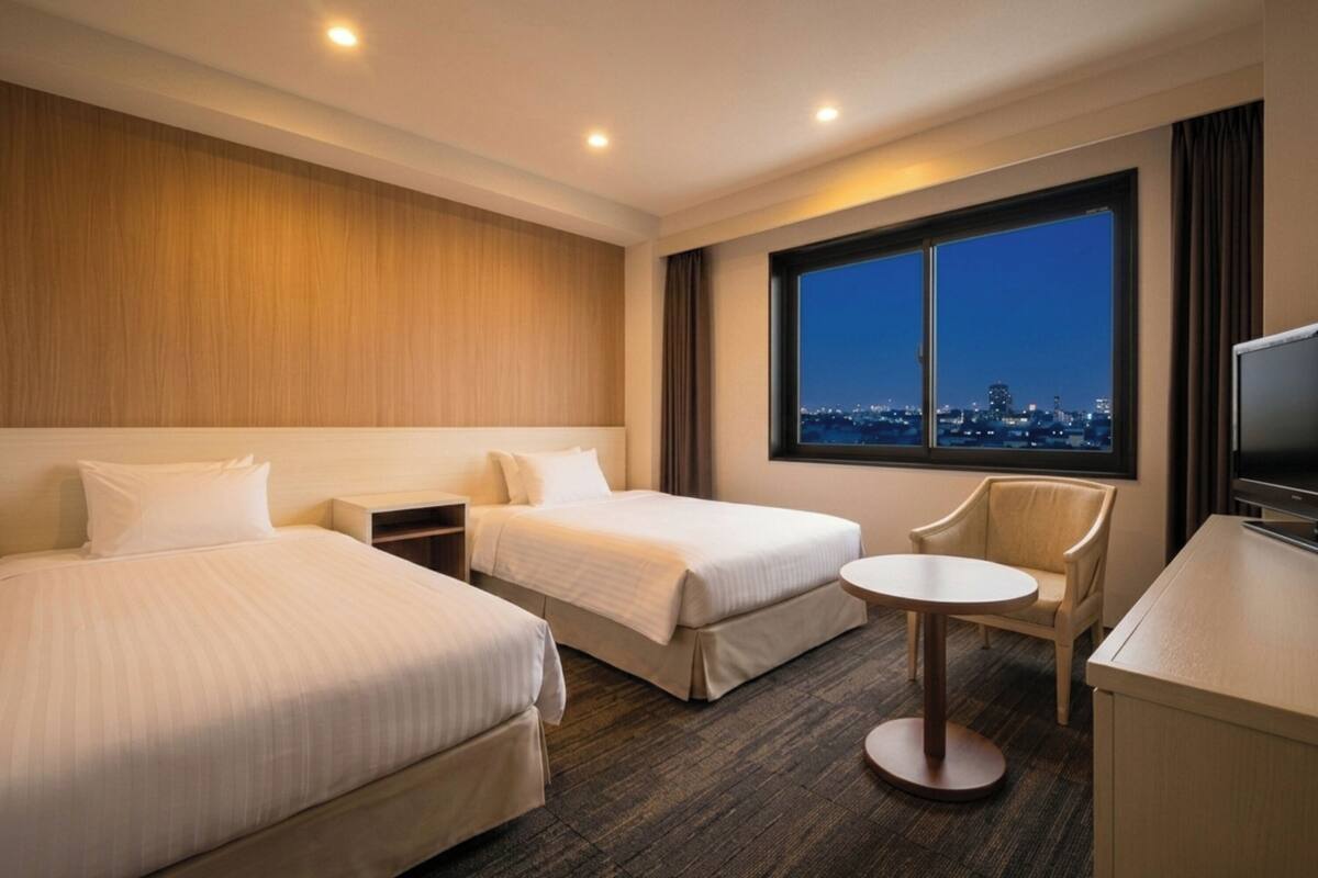 A hotel room with two beds, a small round table, a chair, a TV, and a large window with a city view.