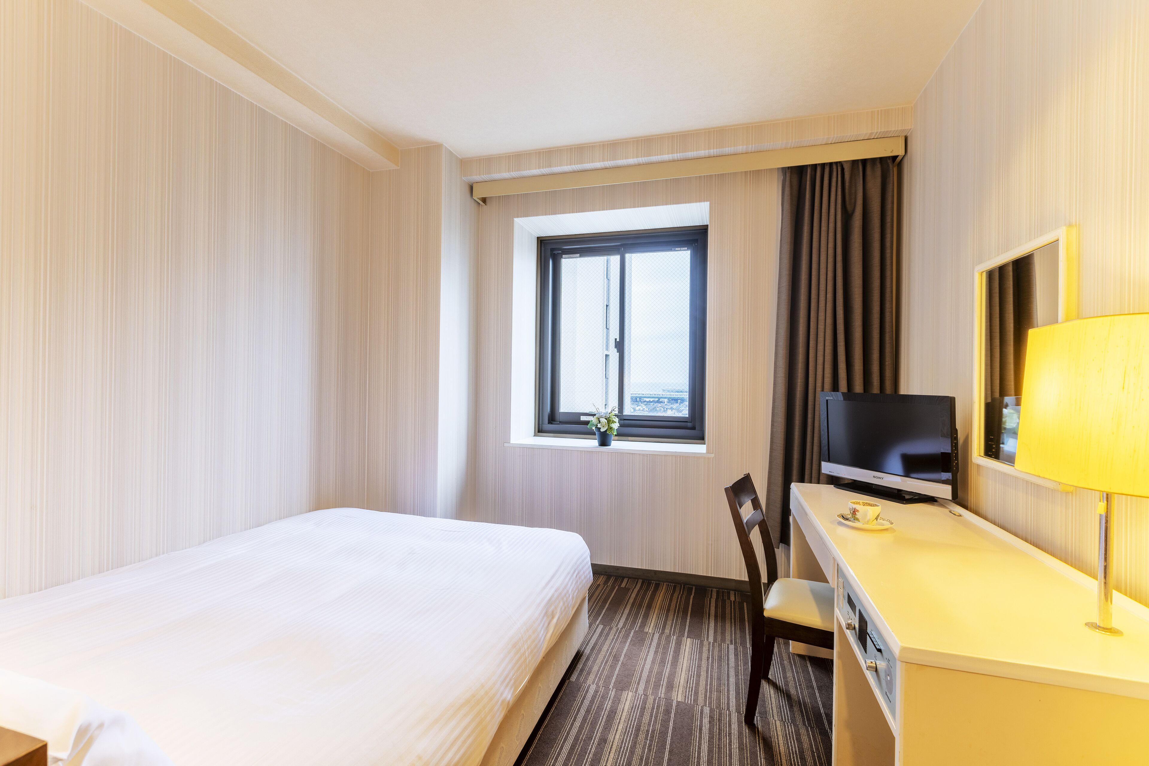 A hotel room with a bed, a desk with a television, a chair, and a window with curtains.