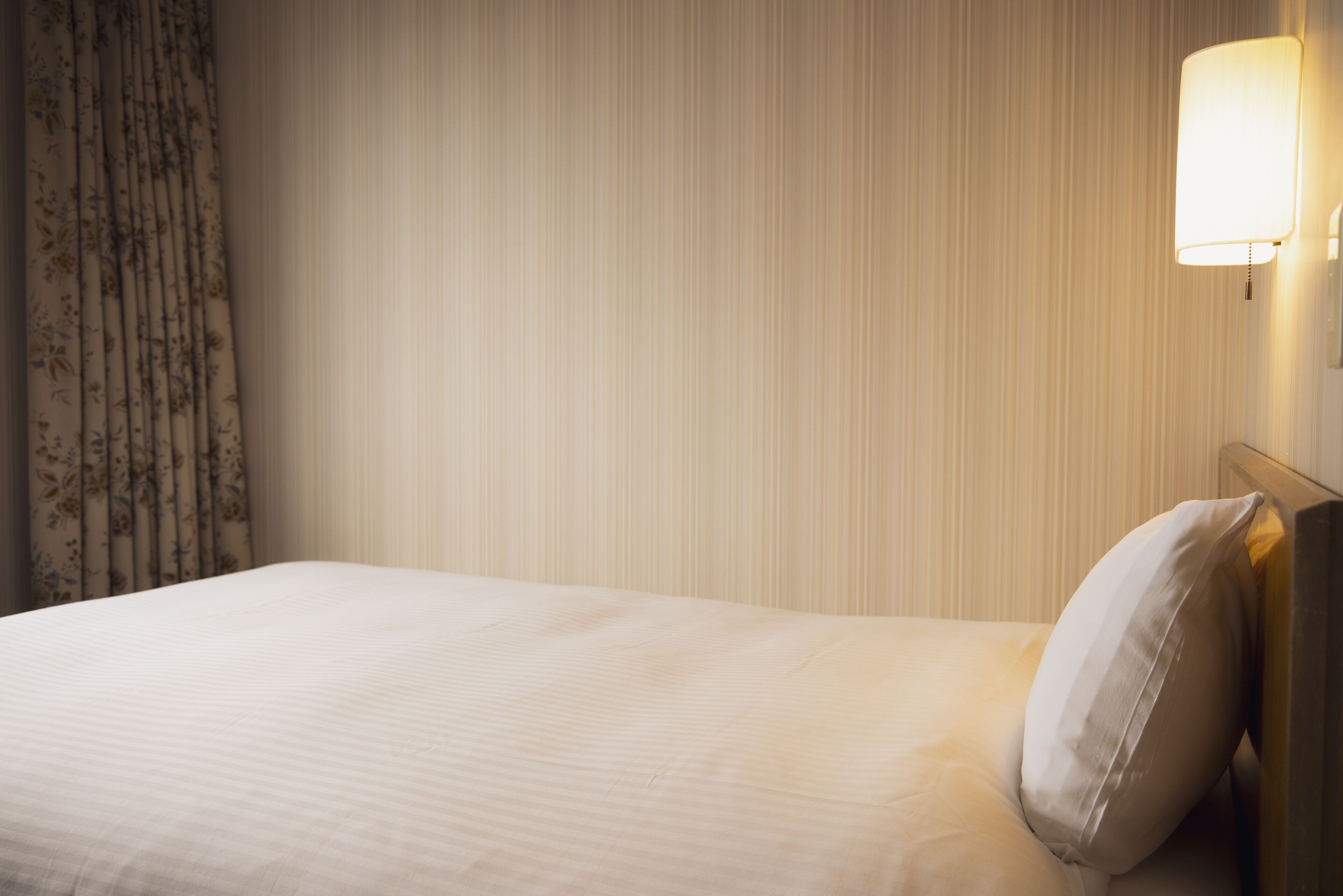 Una cama bien tendida con una almohada, un cortinero floral y una lámpara fijada a la pared.