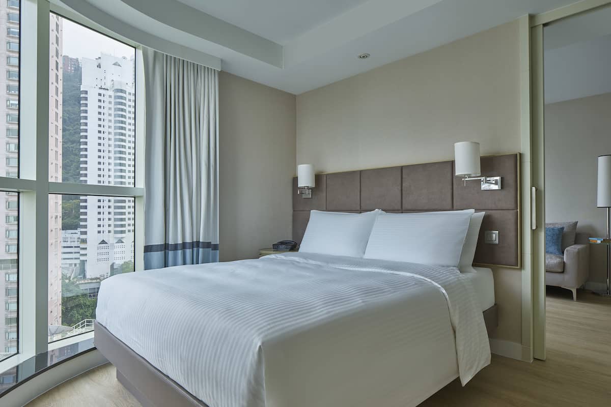 A modern hotel room with a large bed, a nightstand, a lamp, and a view of tall buildings through the window.