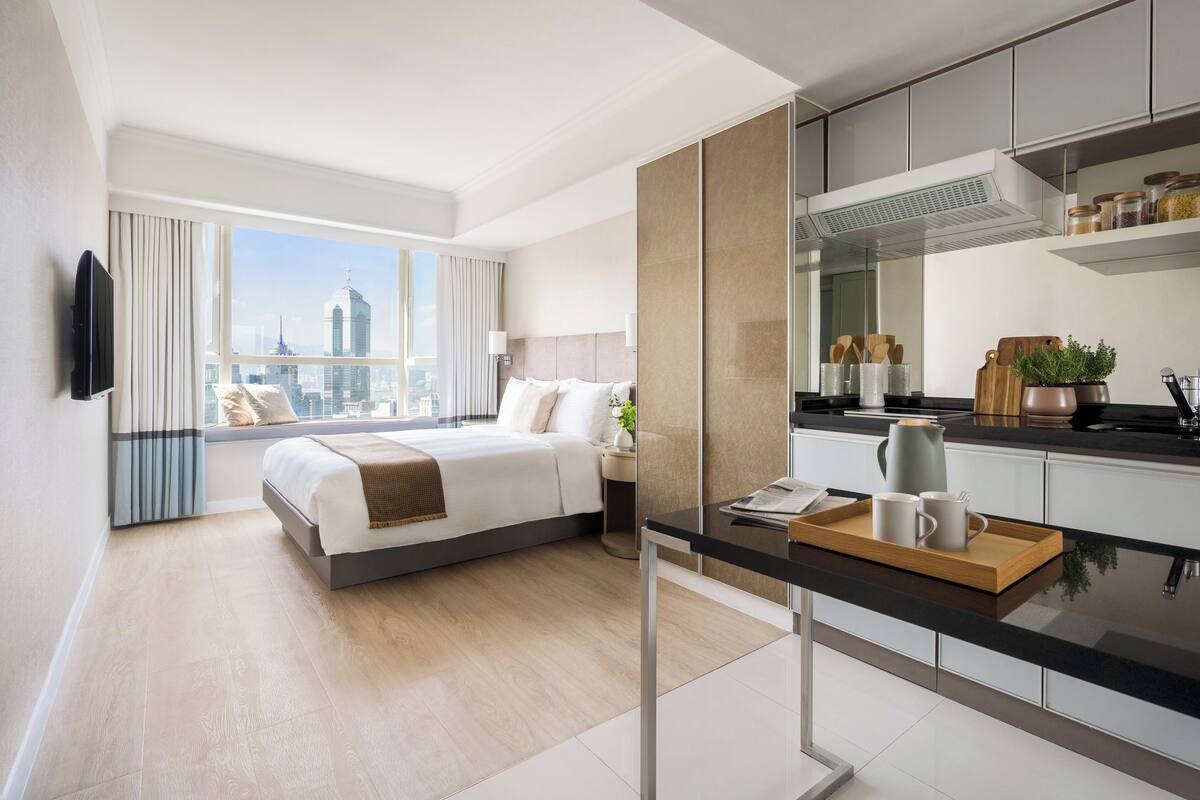 A modern hotel room with a large bed, a dining area, and a view of the city skyline.