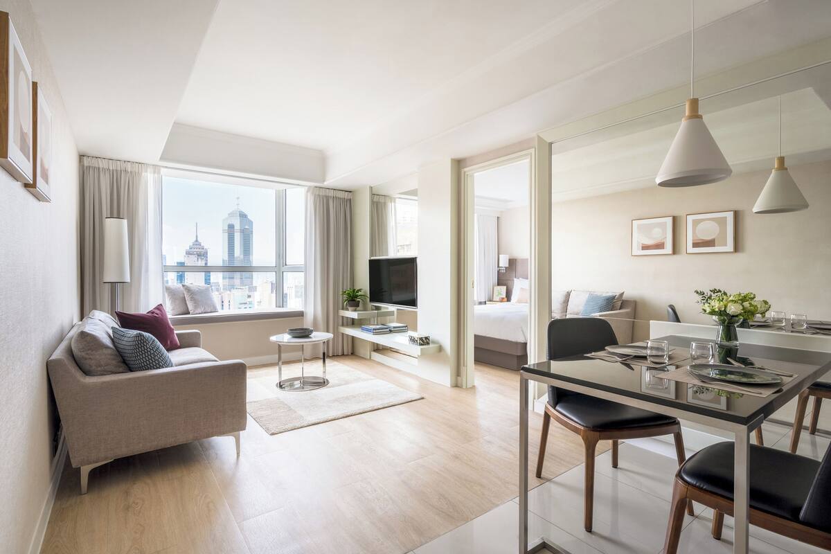 A modern hotel room with a sofa, dining table, and city view.