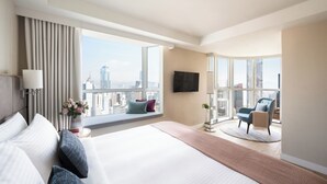 Ein modernes Hotelzimmer mit einem großen Bett, einer Sitzecke mit einem Sessel, einem an der Wand montierten Fernseher und Blick auf die Stadt durch große Fenster.