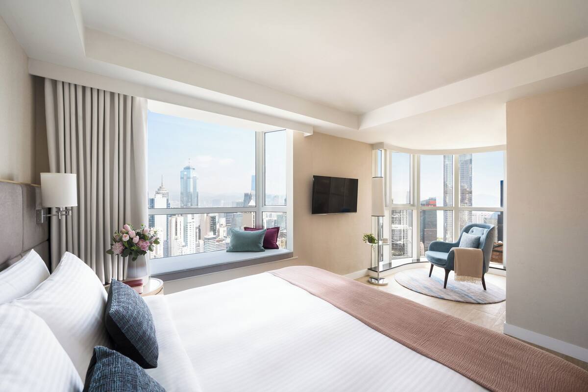 A modern hotel room with a large bed, a seating area with a chair, a TV mounted on the wall, and a view of the cityscape through large windows.