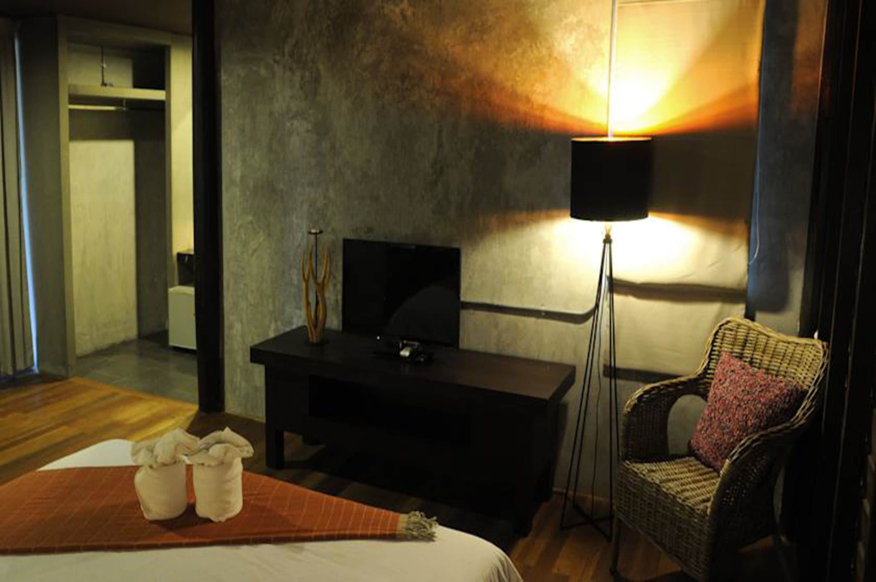 A hotel room with a bed, a television on a black stand, a floor lamp, a wicker chair, and a small table with two white objects on it.