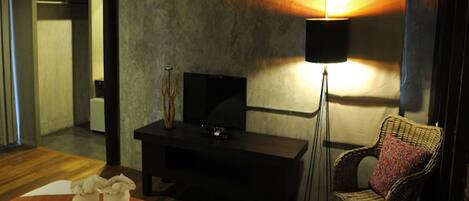 A hotel room with a bed, a television on a black stand, a floor lamp, a wicker chair, and a small table with two white objects on it.
