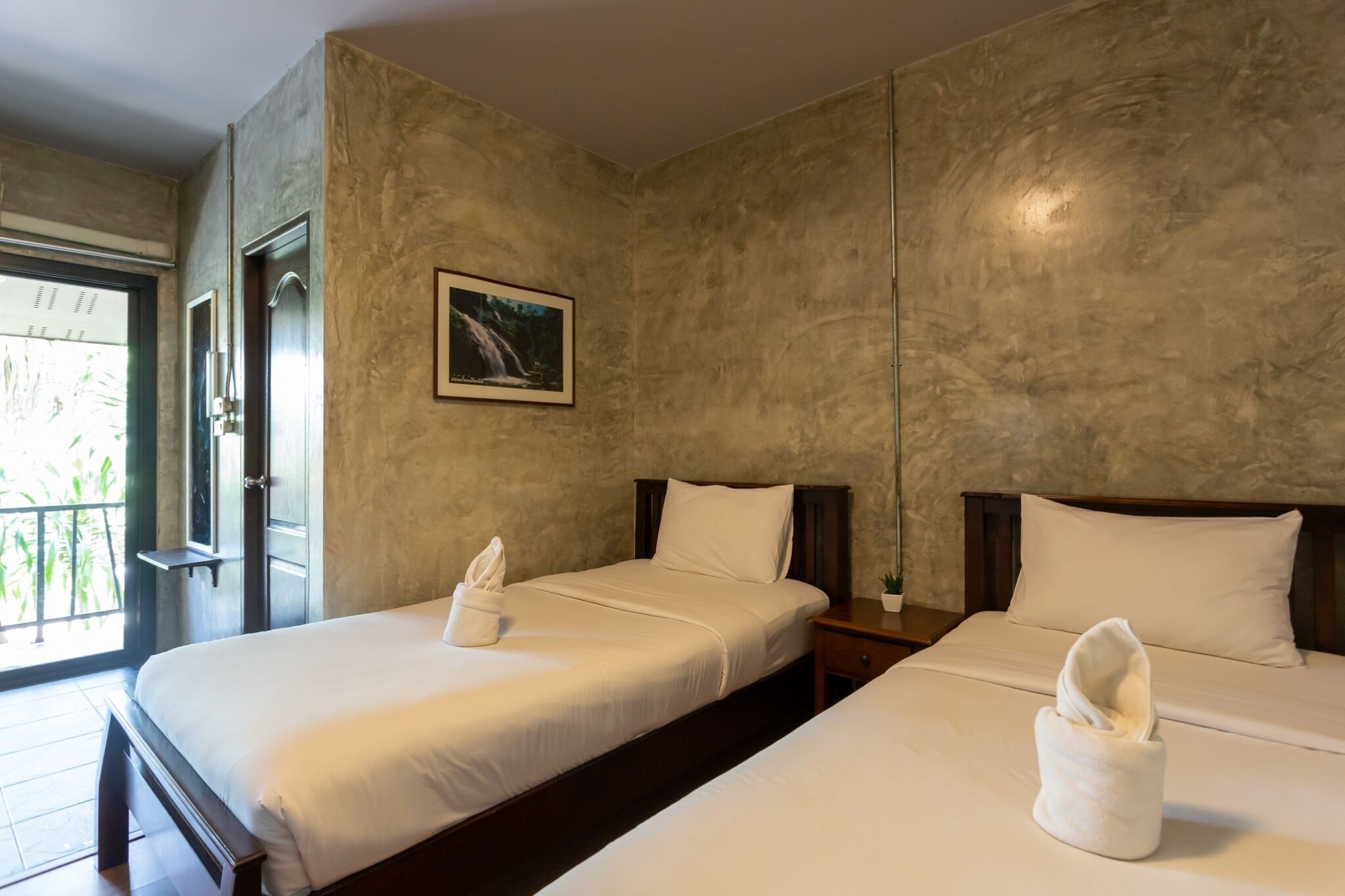 A hotel room with two beds, a framed picture on the wall, and a window with a view of greenery.