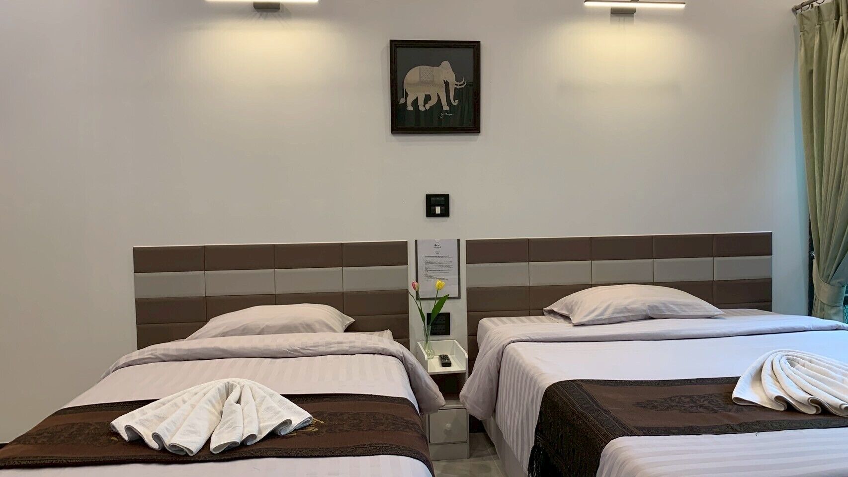 A hotel room with two beds, a framed elephant artwork on the wall, and a nightstand with a vase and a remote control.
