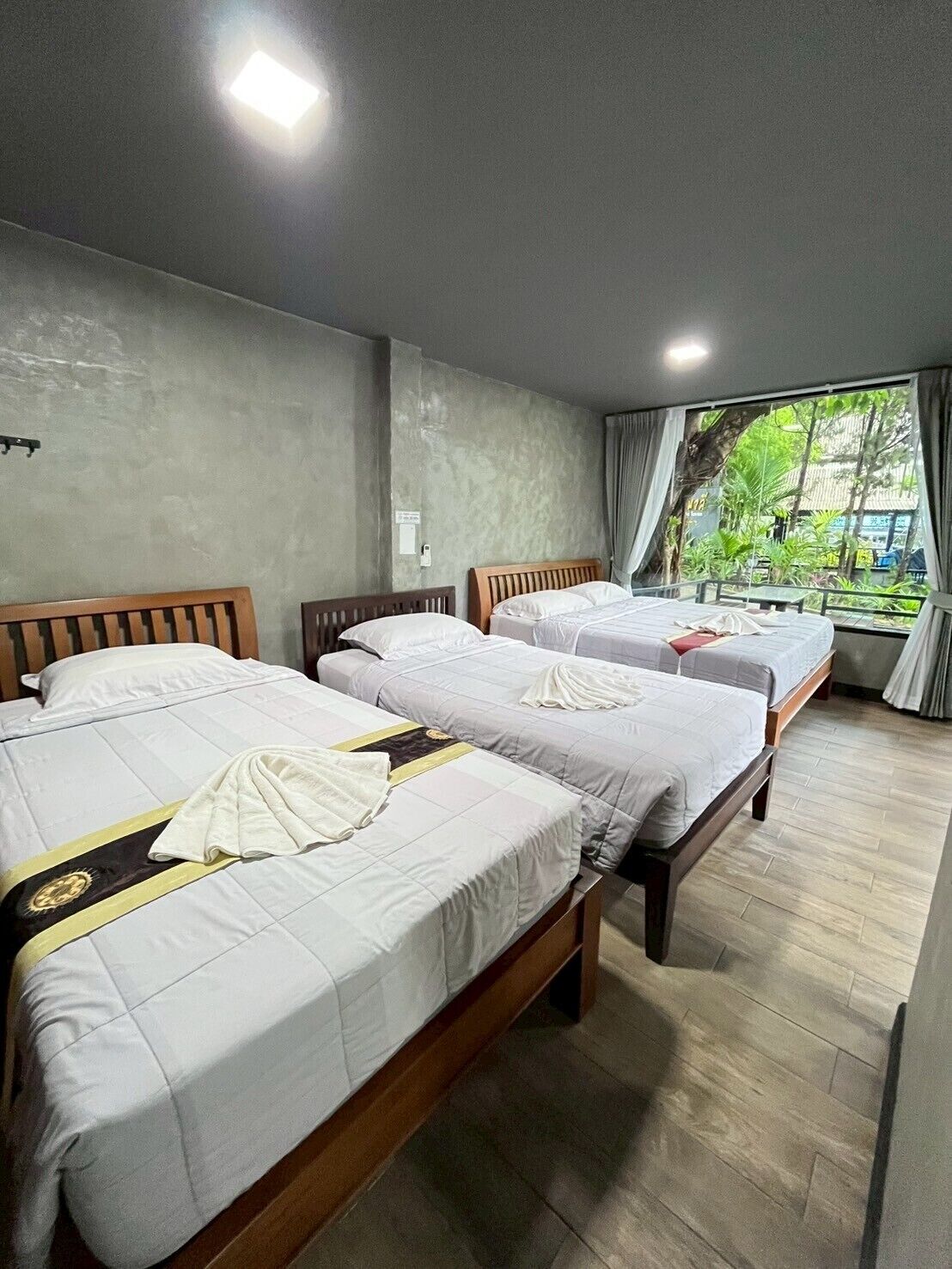 A hotel room with three beds, a large window with curtains, and a concrete wall.