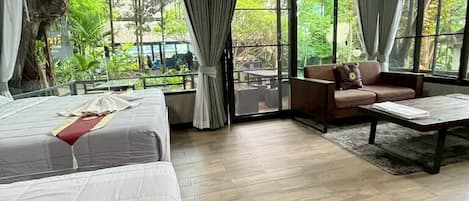 A spacious room with a large bed, a sofa, and a coffee table. There are large windows with curtains, and a view of greenery outside.