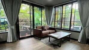 Una habitación amplia con piso de madera, un sofá marrón, una mesa de centro y ventanas grandes con cortinas.