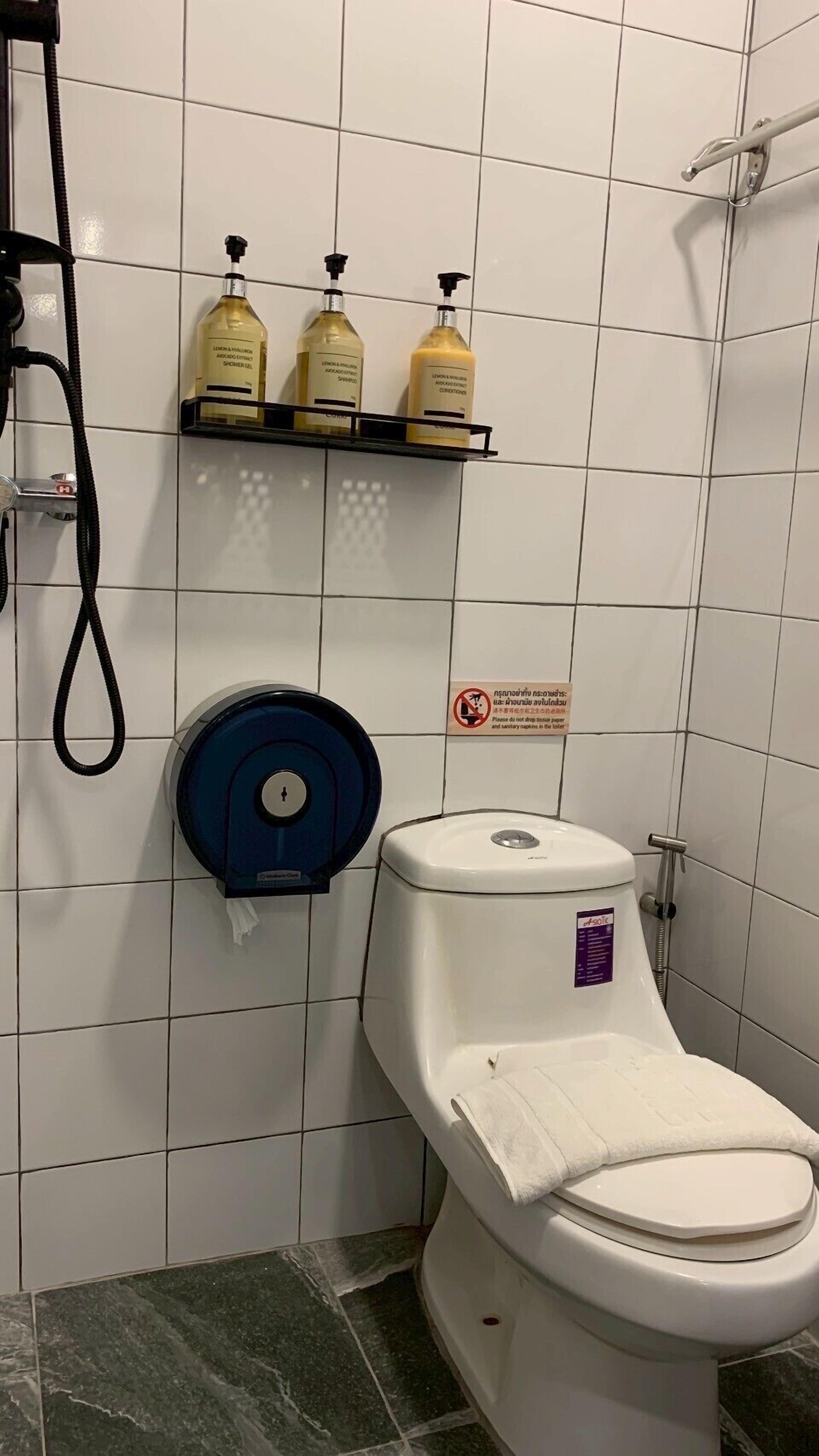 Un baño con un inodoro, un secamanos azul y tres botellas de gel de ducha en un estante.