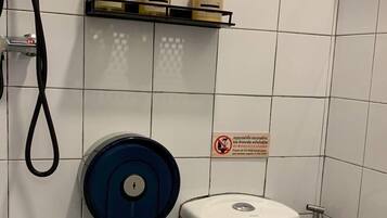 Un baño con un inodoro, un secamanos azul y tres botellas de gel de ducha en un estante.