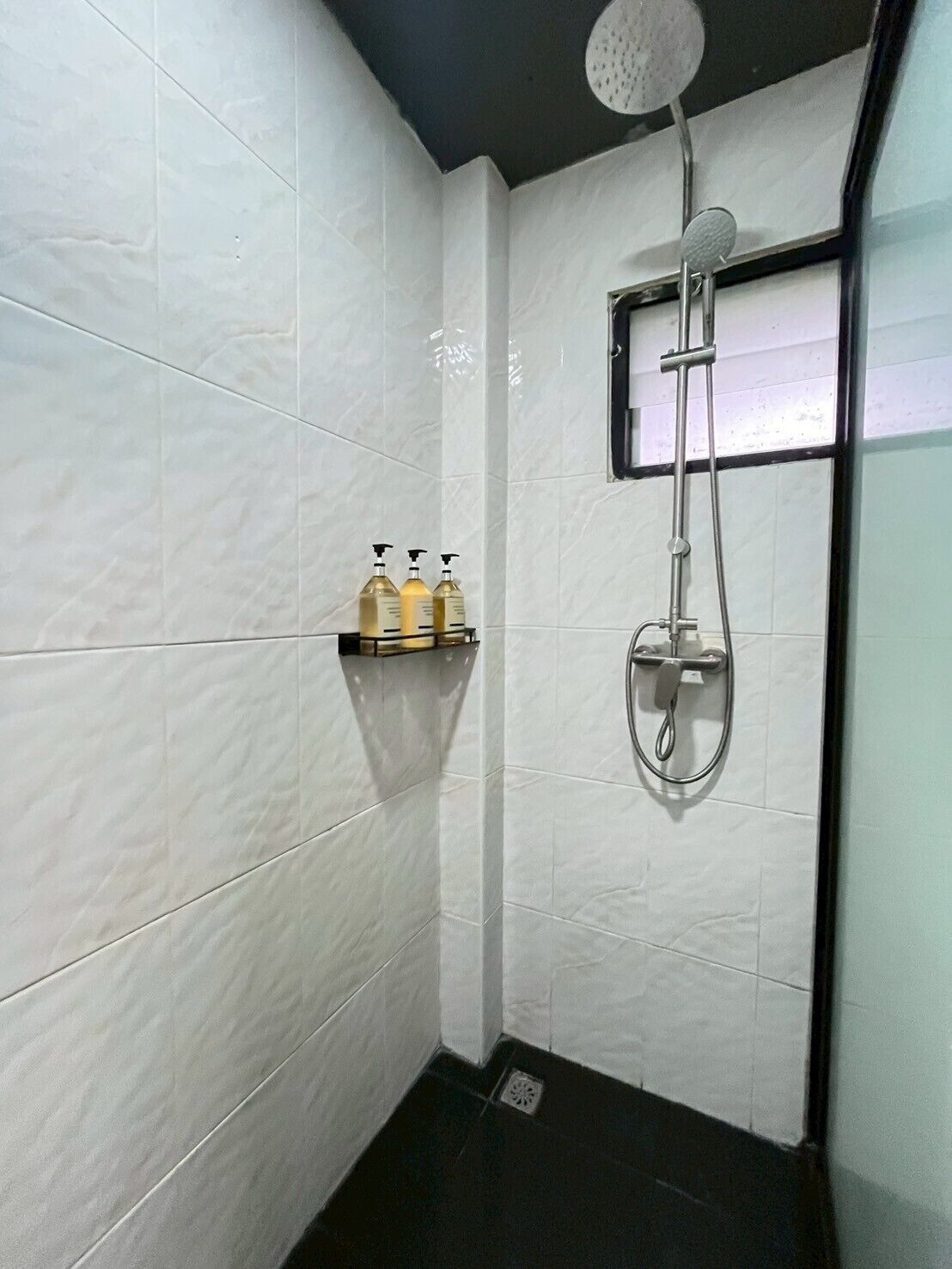 Un baño pequeño y moderno con azulejos blancos, una ducha con cabezal de ducha manual y un estante con tres botellas.