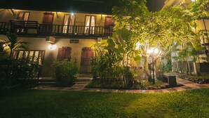 Un patio de hotel por la noche con un jardín iluminado, una piscina y un edificio de dos plantas con balcones.