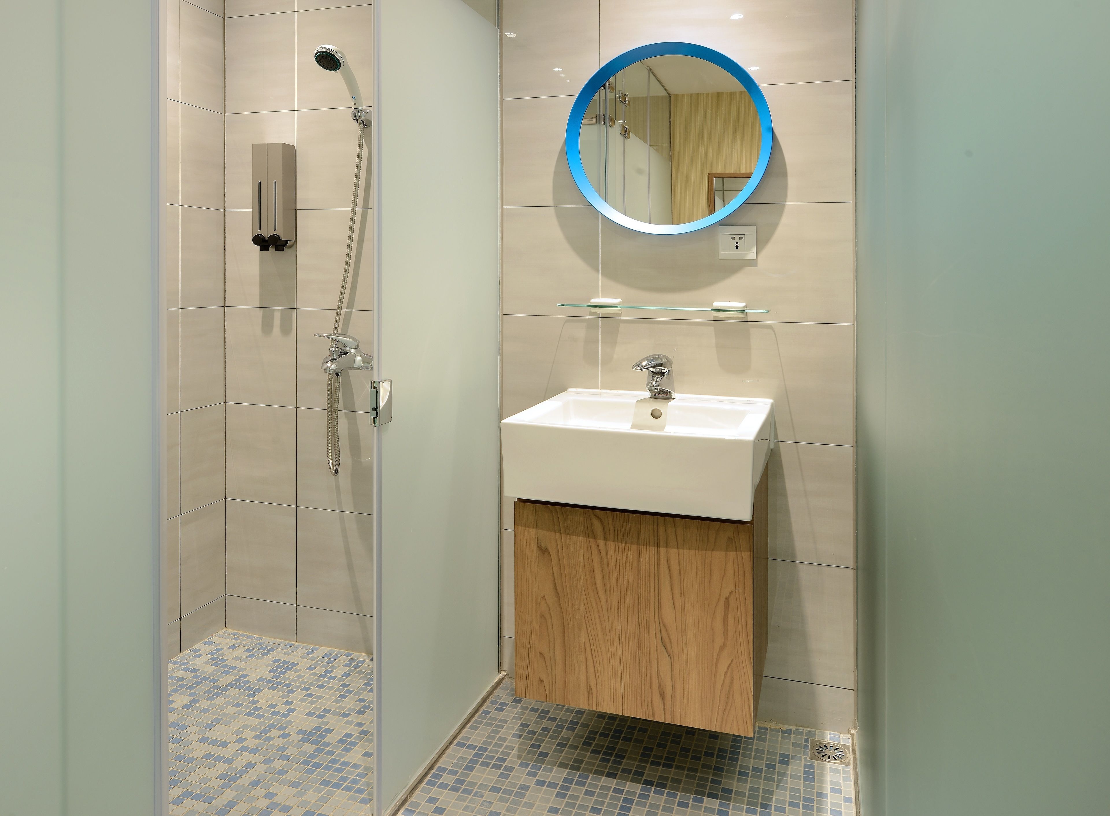 A modern bathroom with a glass-enclosed shower, a white sink with a wooden vanity, and a round mirror with a blue frame.