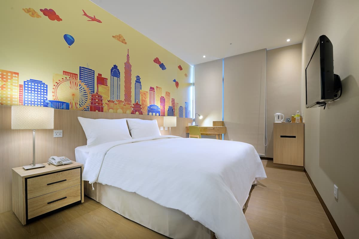 Een hotelkamer met een bed, nachtkastje, lamp en een stadsgezichtsmural op de muur.