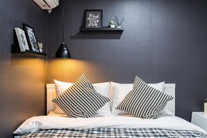 Un letto rifatto con lenzuola bianche e cuscini a motivi bianchi e neri, una lampada a pendente nera e uno scaffale con quadri incorniciati e una piccola pianta.