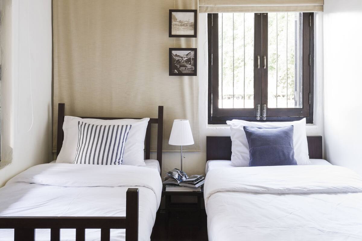 Una habitación con dos camas, una ventana con persianas, una mesita de noche con lámpara y un cuadro enmarcado en la pared.