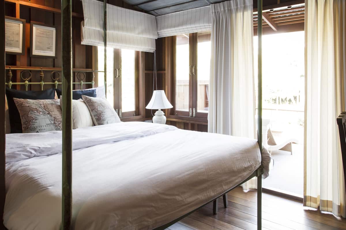 Una cama grande con cabecera de madera, una mesita de noche con lámpara, y vista al exterior a través de una ventana con cortinas.