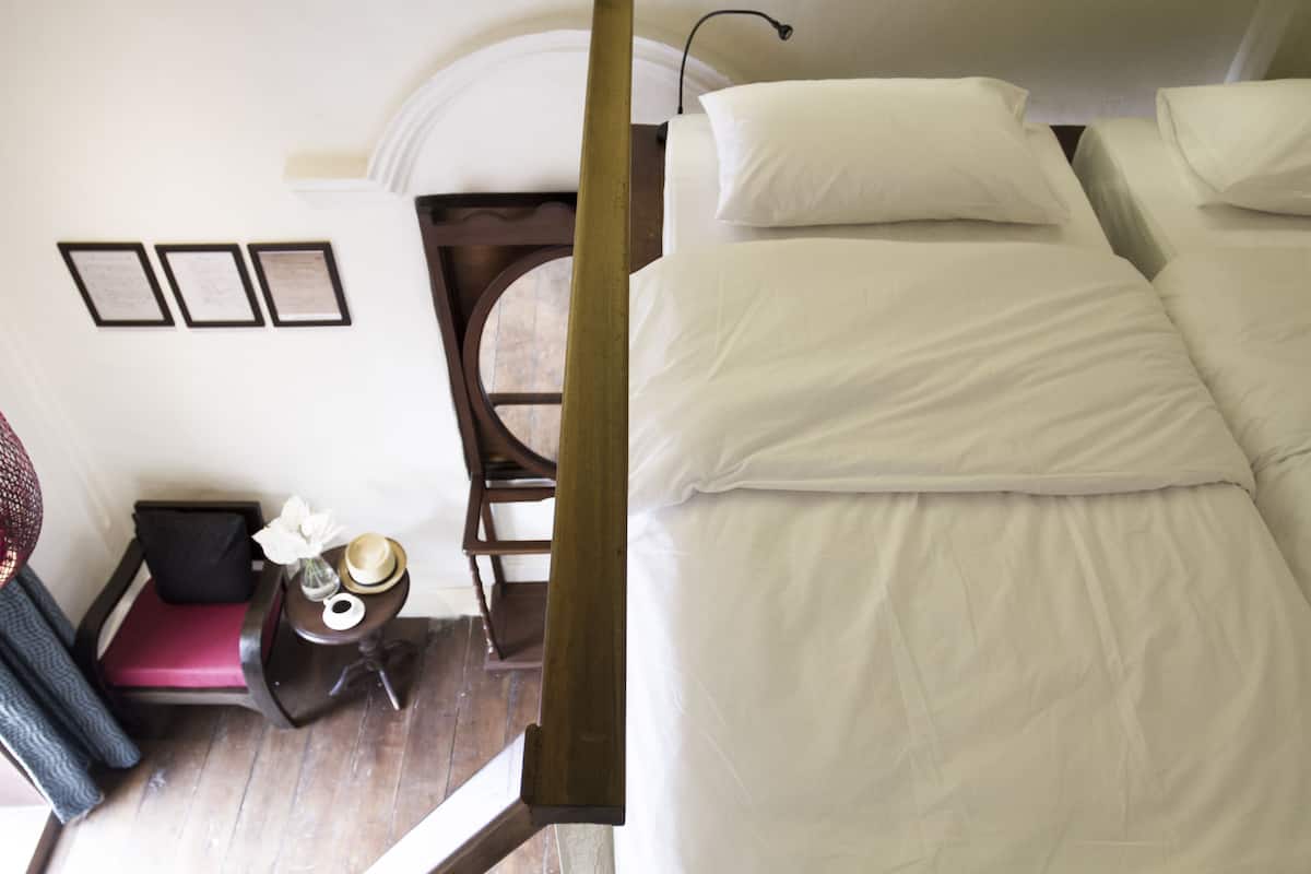 Una habitación de hotel con una cama grande, una mesita de noche pequeña con un florero y un sombrero, una silla y una ventana con vistas.