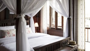 A four-poster bed with a canopy, a bedside table with a lamp, and a window with curtains. - Baan Luang Rajamaitri Historic Inn (Chanthaburi)