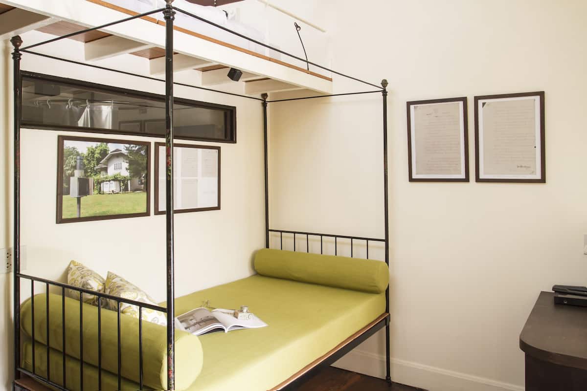 Habitación individual con cama de estructura metálica, ropa de cama verde, ventana con vista a una casa y dos documentos enmarcados en la pared.