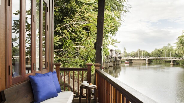 A balcony with a wooden bench, a small table with a cup and a pastry, and a view of a river with a bridge in the background. - Baan Luang Rajamaitri Historic Inn (Chanthaburi)