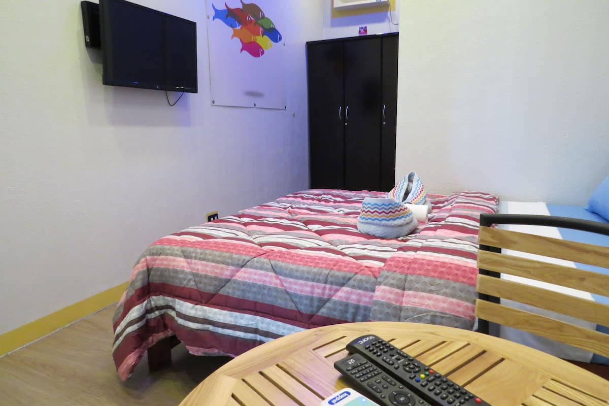 Una habitación con cama, una silla de madera, una mesa con controles remotos, un televisor en la pared y un armario.