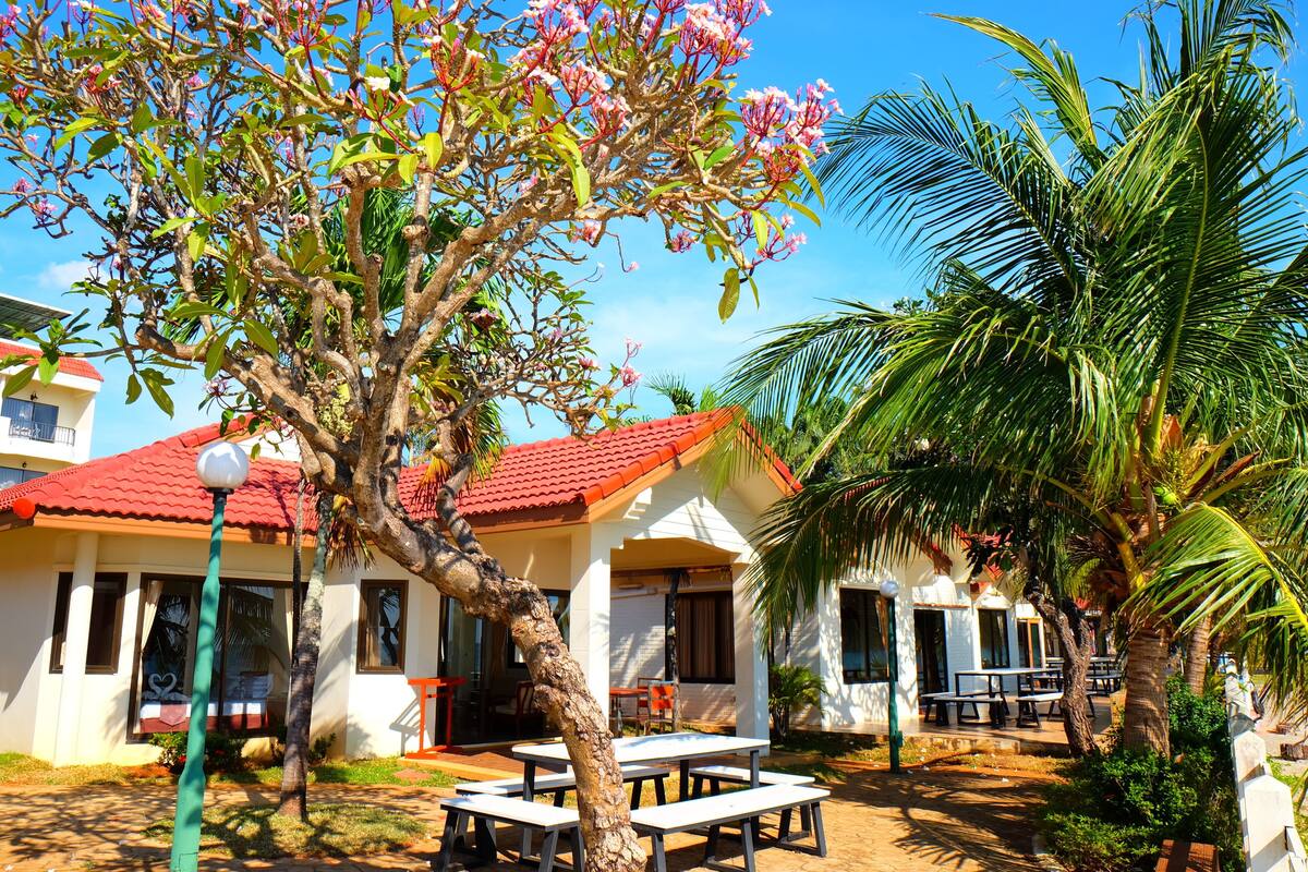 Un bungalow avec un toit recouvert de tuiles rouges, un espace salon extérieur et un arbre aux fleurs roses.