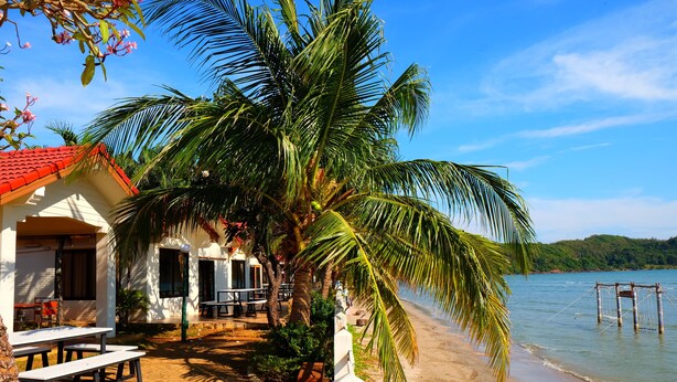 A beachfront property with a red-roofed building, outdoor seating, and a palm tree. - New Travel Beach Resort (Tha Mai)