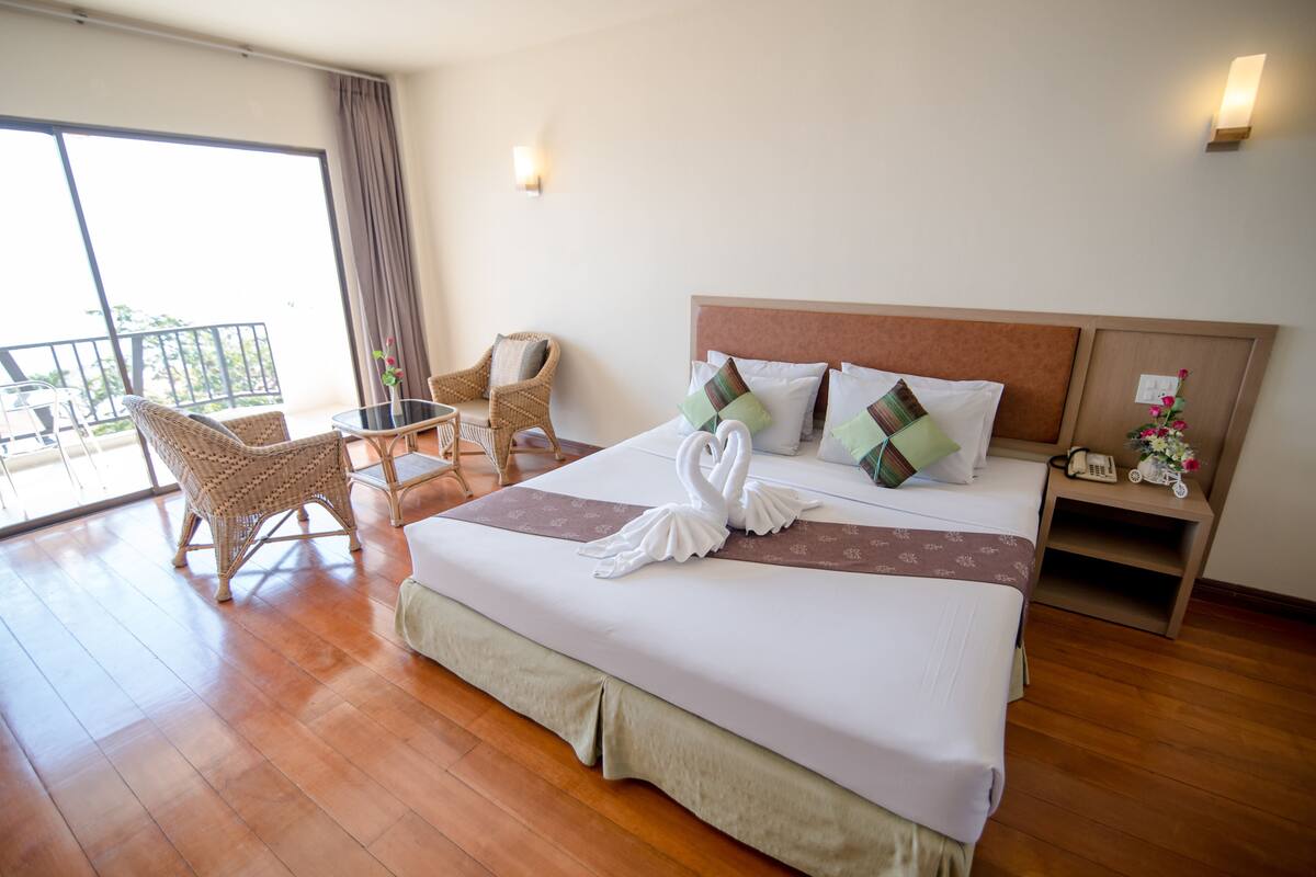 Une chambre d’hôtel avec un grand lit, un balcon, des fauteuils en osier, une petite table et une table de chevet avec un téléphone et des fleurs.