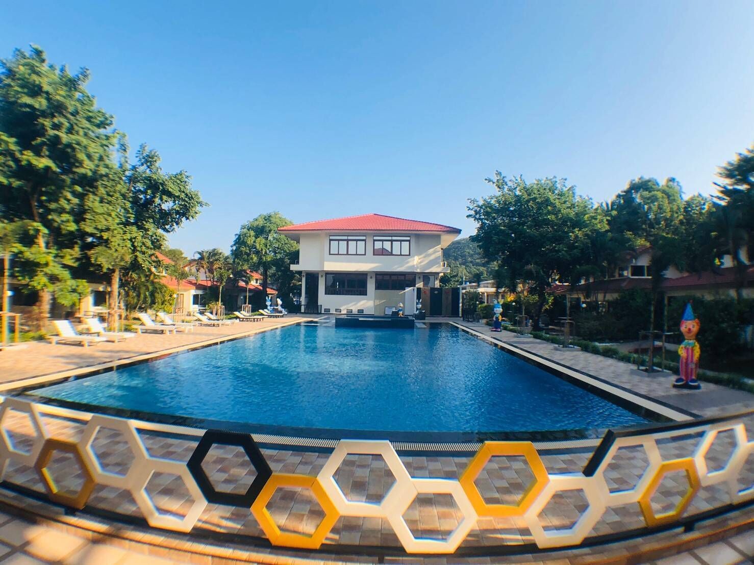 Una piscina con borde geométrico, un edificio de techo rojo y sillas de descanso alrededor de la piscina.