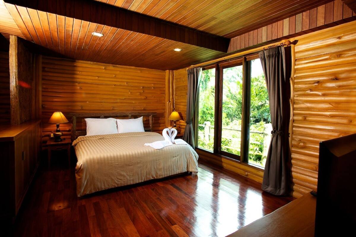 A log cabin-style bedroom with a wooden bed, two bedside lamps, a window with curtains, and a view of greenery outside.