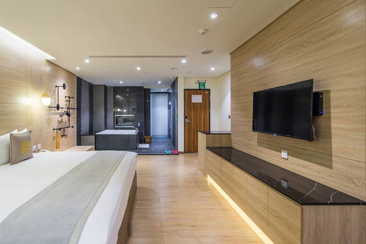A modern hotel room with a large bed, a flat-screen TV mounted on the wall, and a wooden floor.