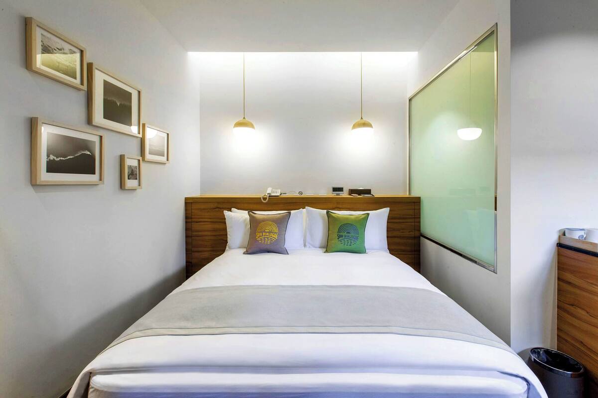 A hotel room with a neatly made bed, a wooden headboard, and a large window. There are framed pictures on the wall and a small desk with a phone and a lamp.