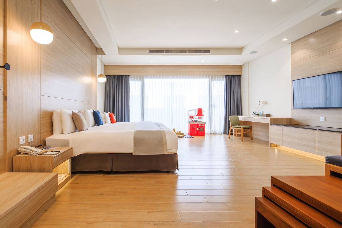 A modern hotel room with a wooden floor, a bed with multiple pillows, a desk with a chair, a TV mounted on the wall, and a large mirror.