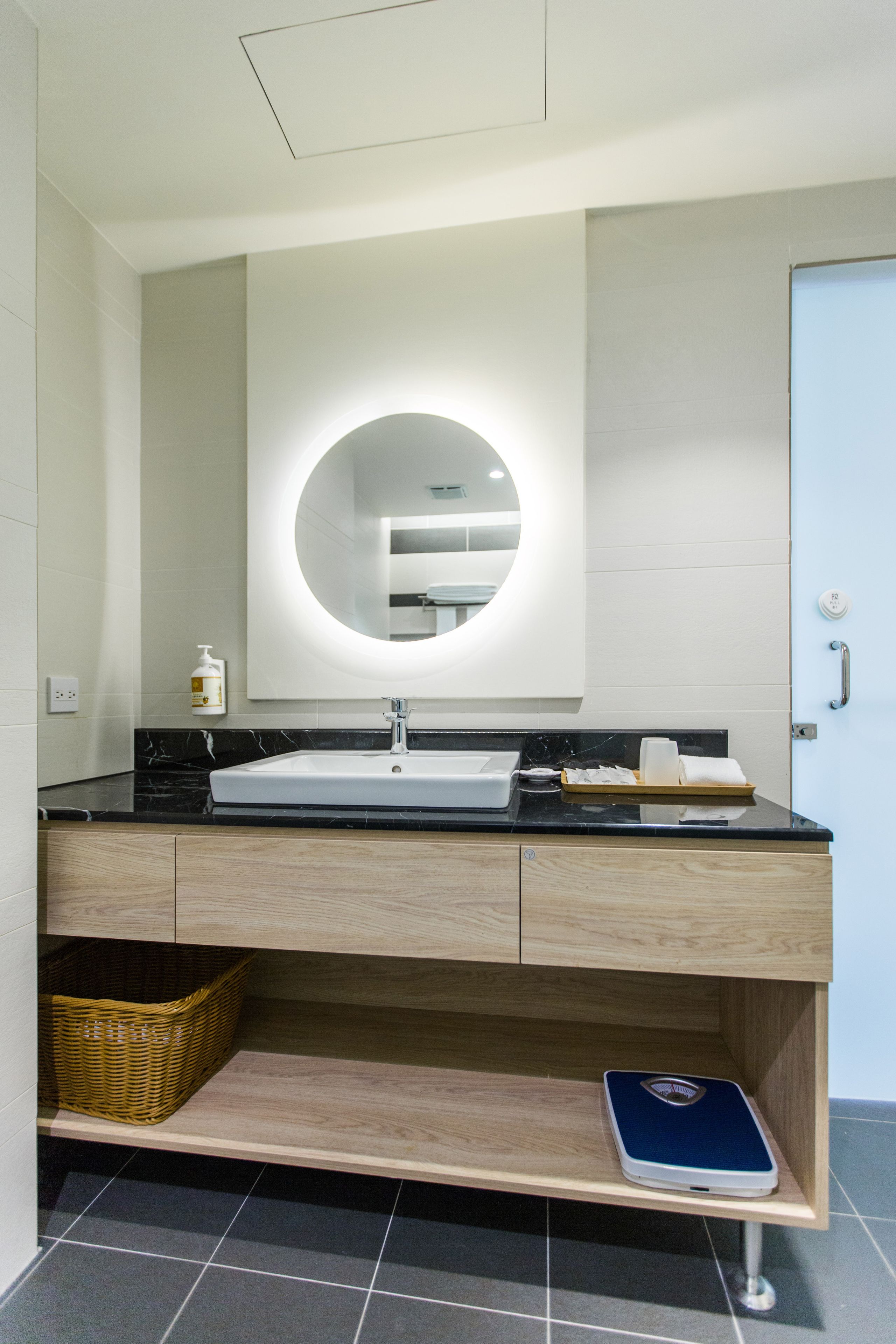 Un baño moderno con un lavamanos blanco, encimera negra, mueble de tocador de madera y una balanza en el estante inferior.