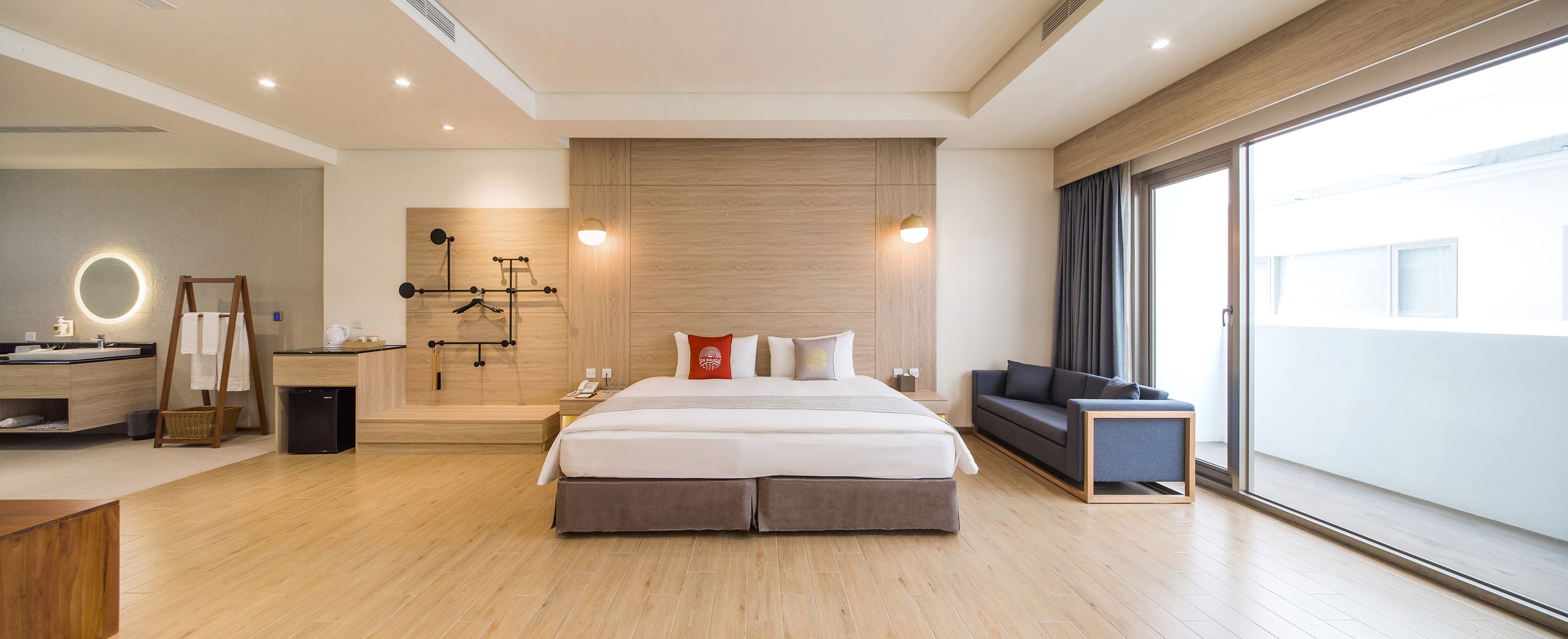 A modern hotel room with a large bed, a sofa, a vanity with a mirror, and a view of the city through large windows.