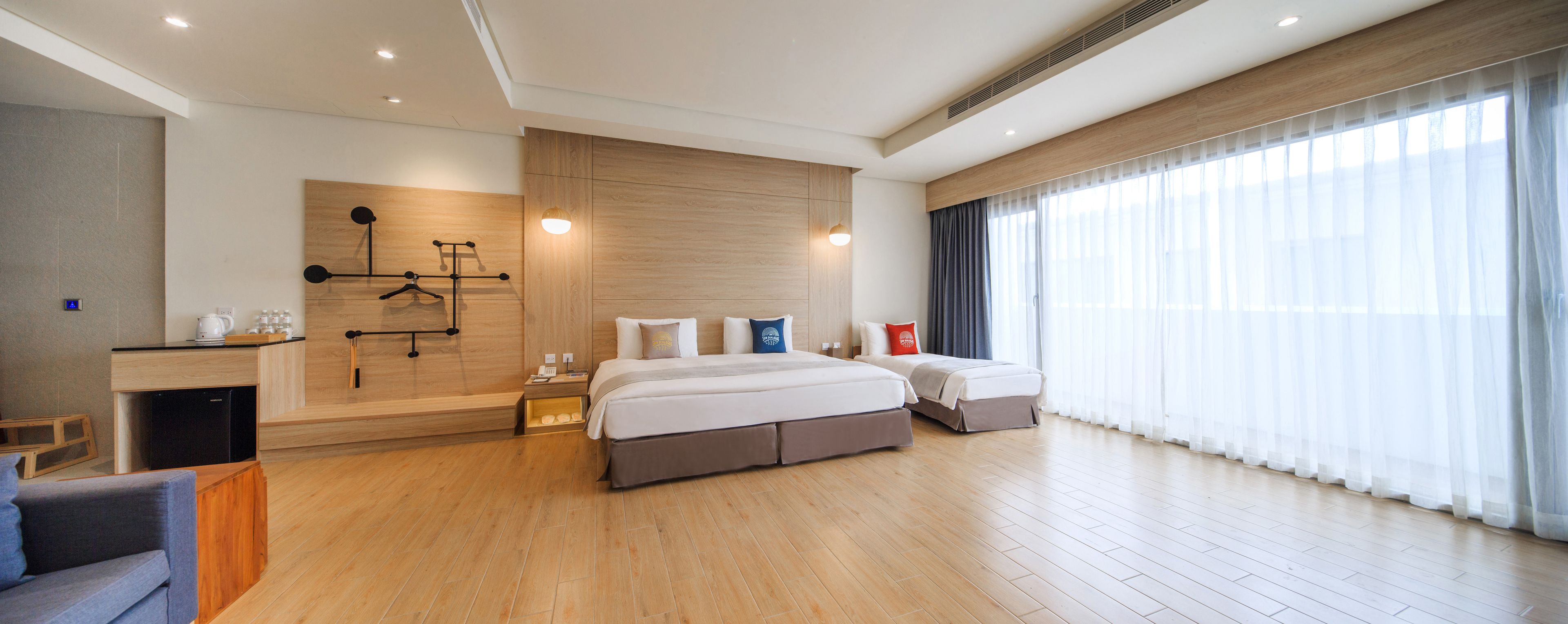 Een moderne hotelkamer met een groot bed, een bureau met koffiezetapparaat, een kledingkast en een groot raam met dunne gordijnen.