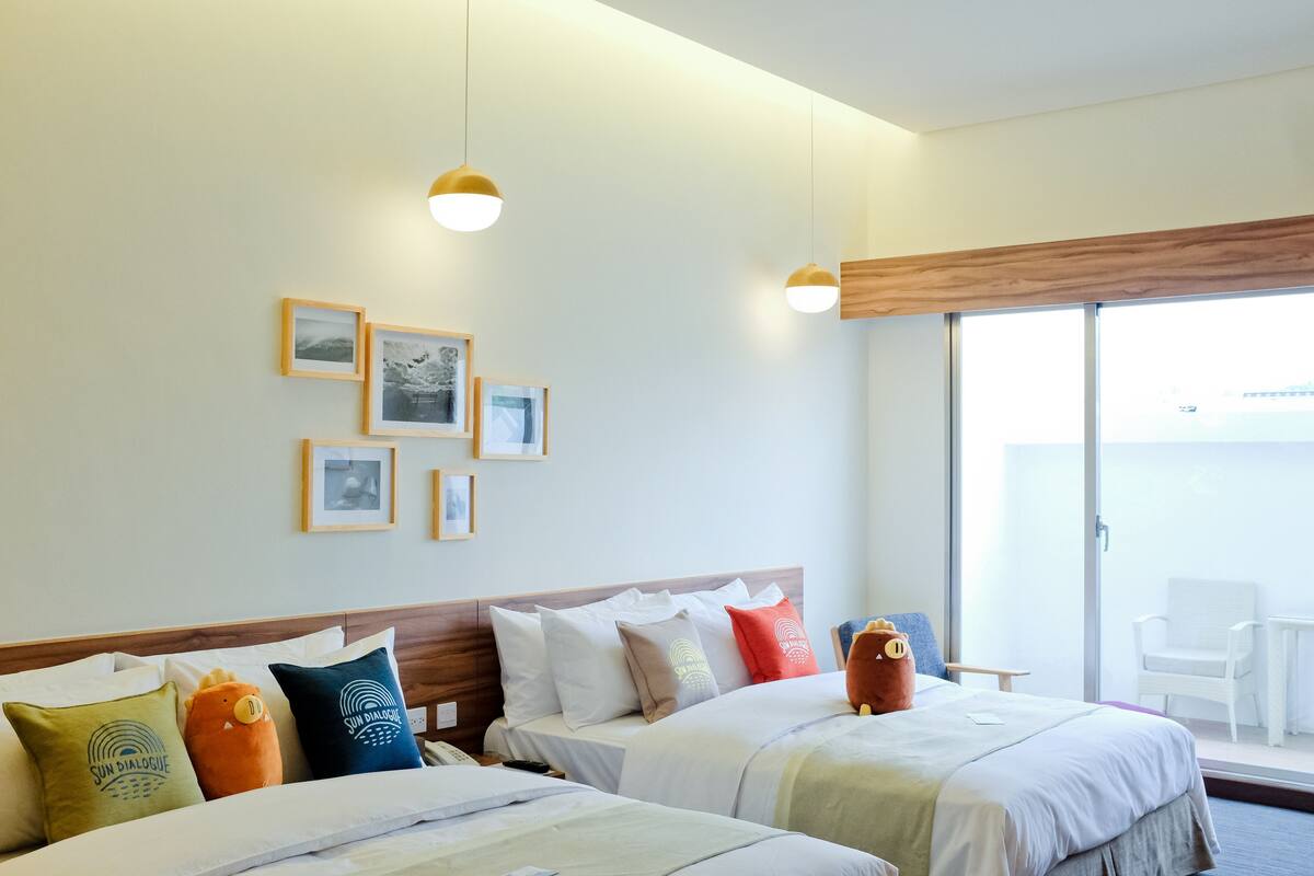 A hotel room with two beds, a wooden headboard, a large window, and a balcony with a white chair.
